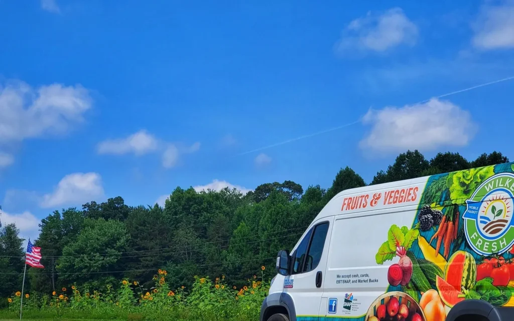A delivery van branded with "Fruits & Veggies" parked near a field of sunflowers, under a bright blue sky with fluffy clouds.